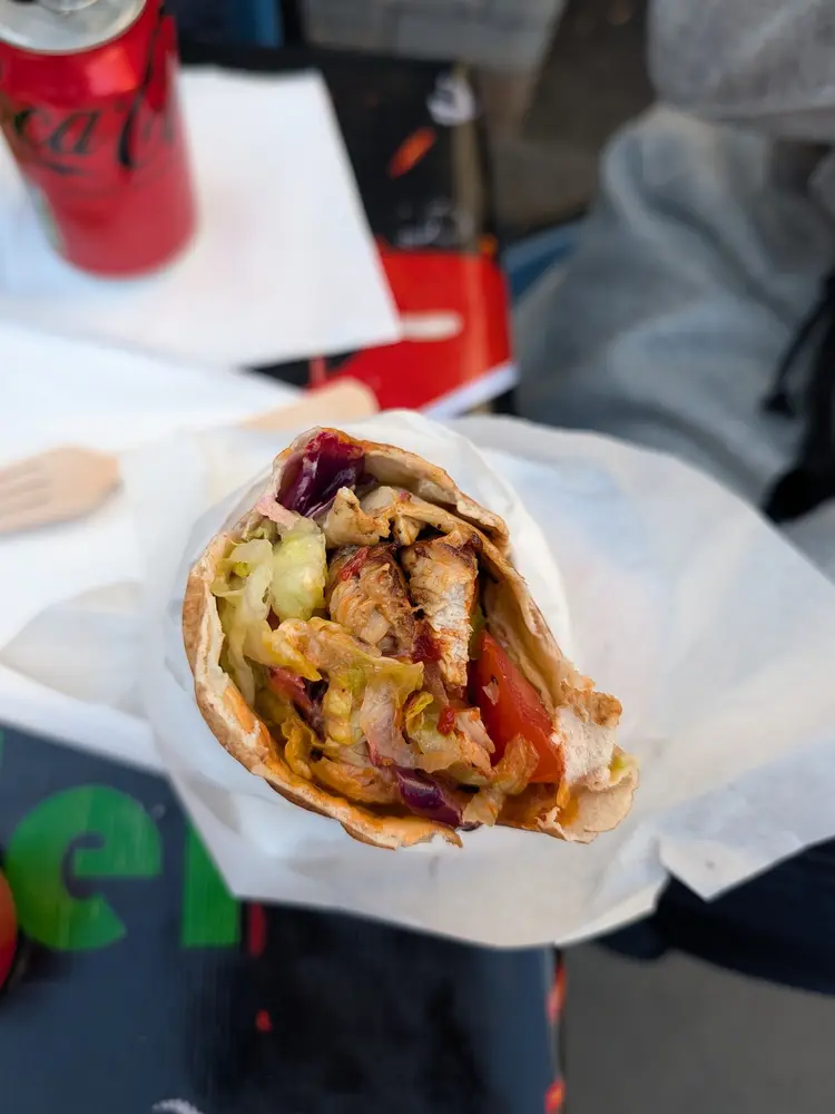 Hand holding an open chicken shish wrap with lettuce, tomato and red cabbage in paper